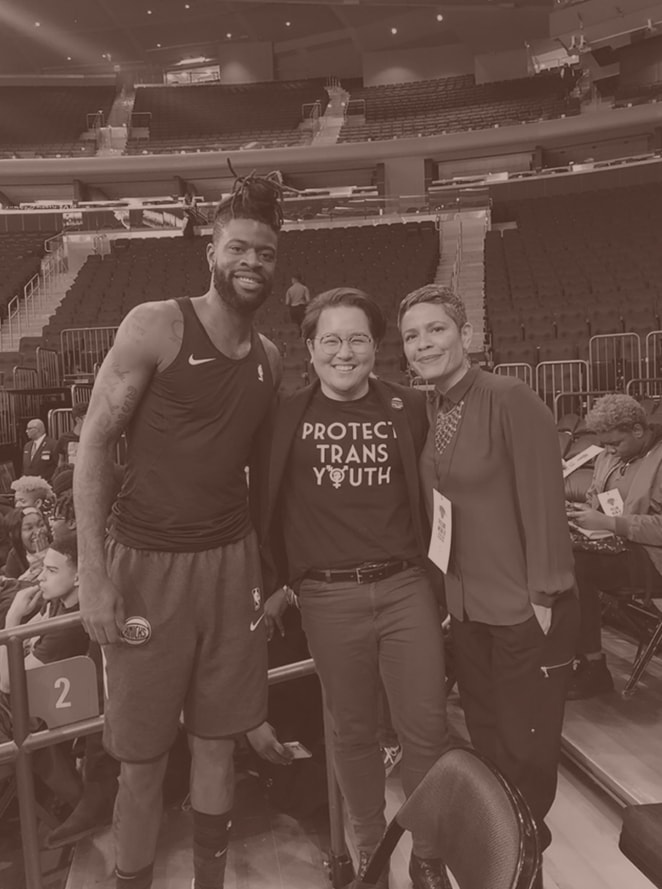 Three People stand together with arms wrapped around each other smiling at a sporting arena, the person in the center, a Japanese American, has a t-shirt that reads "Protect Trans Youth"