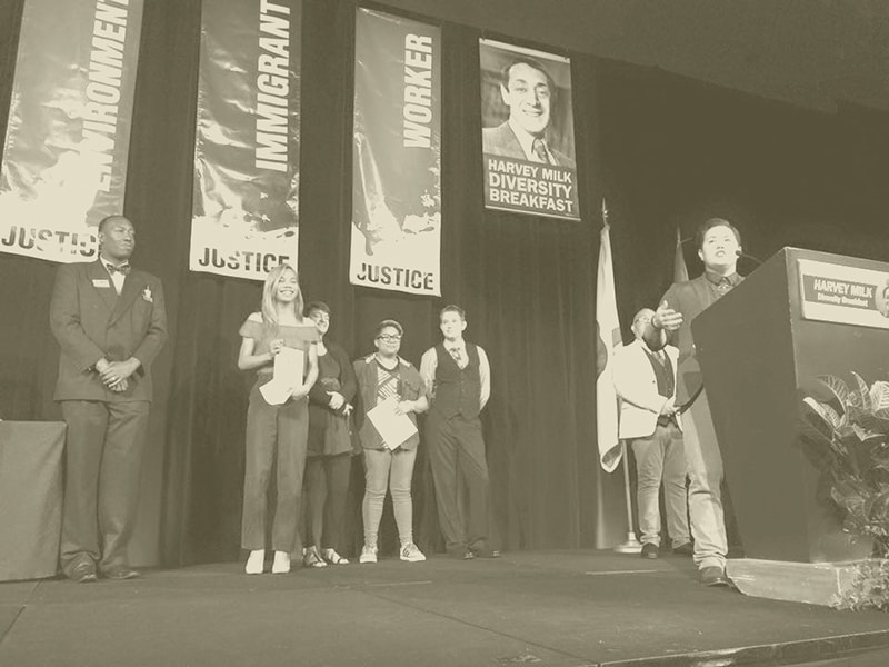At a podium, on a stage, Japanese American, a.t. Furuya, speaks publicly about Award from the SD LGBT Center, four people stand beside a.t. applauding beneath banners that read "Justice" on issues for the immigrant, worker, and environment