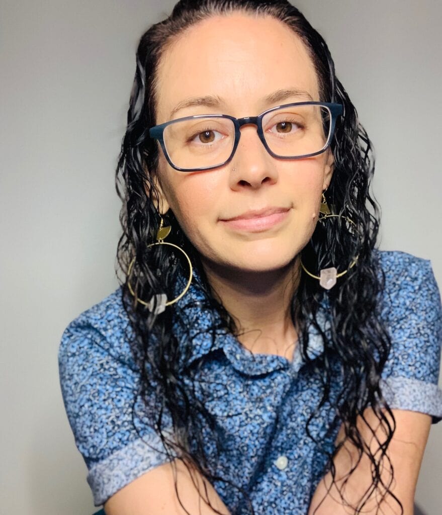 a foto of melissa in a blue flowered button up shirt with rolled up sleeves with a white-grayish backdrop. They have their brown curly hair down. melissa is wearing their teal-indigo colored glasses and dangling gold hoops with rose quartz crystals. End description.